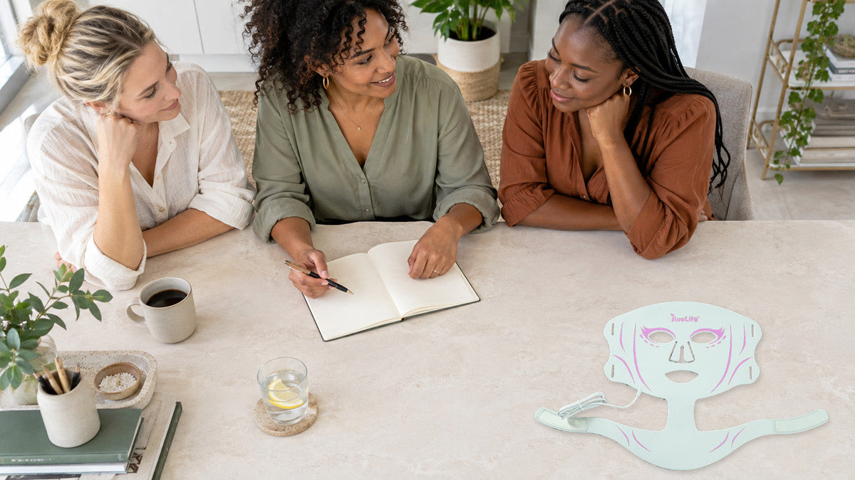 Three adults with different skin tones discussing a notebook beside a white-and-pink face and neck mask on a stone table.