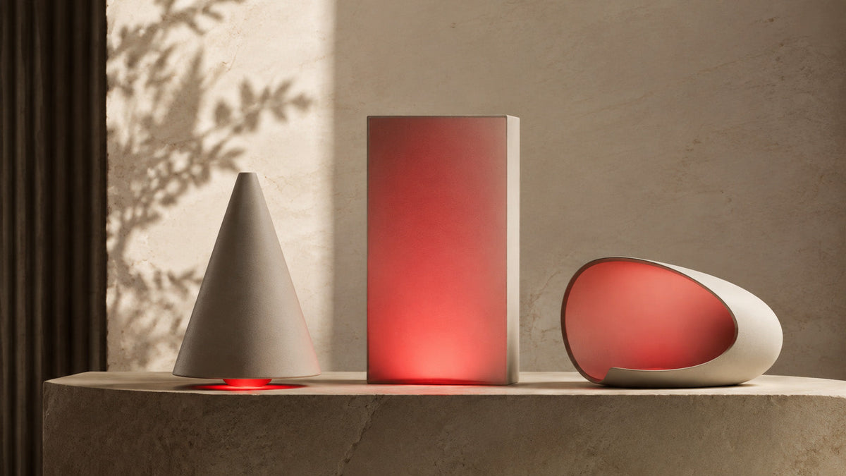 Three abstract ceramic forms, a cone, rectangle and curved oval, illuminated with soft red light on a stone shelf.