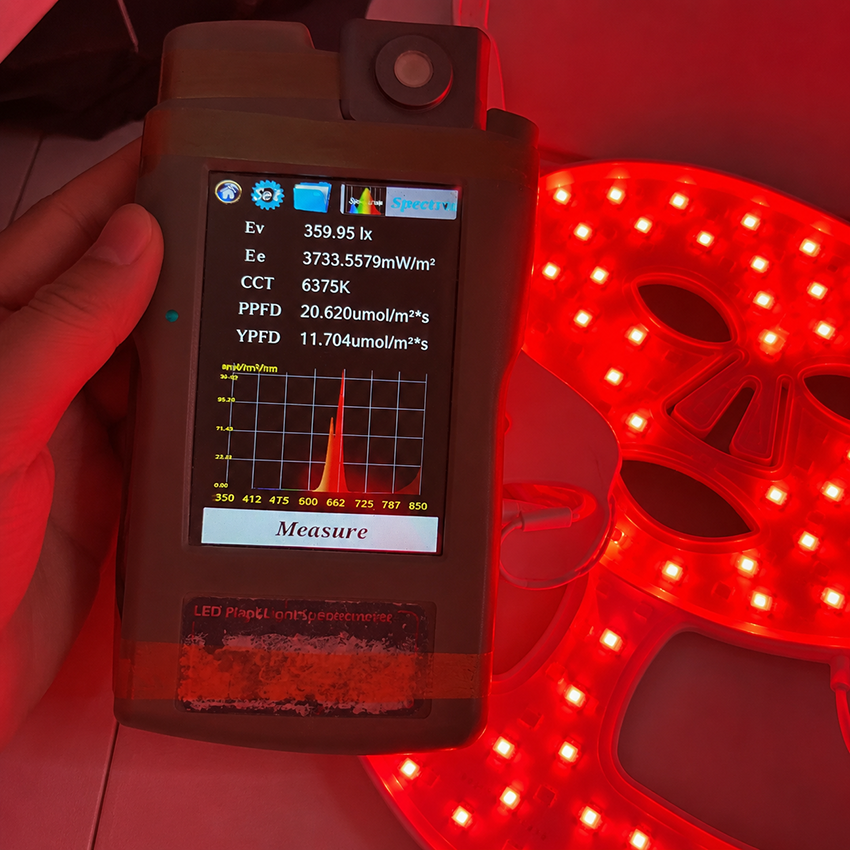 Handheld spectrometer measuring red light output from a glowing LED therapy mask for face and neck care