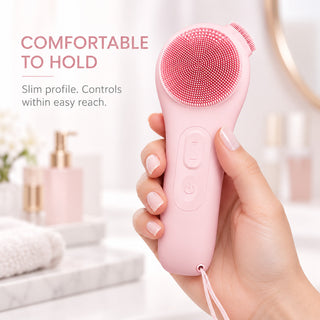 Hand holding the slim pink silicone facial cleansing brush, showing its grip, front controls and wrist strap.